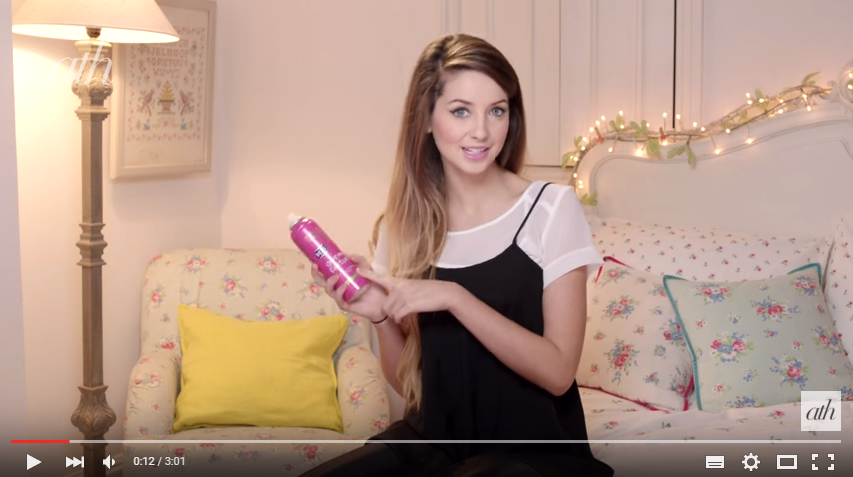 YouTube 明星 Zoella 的鱼尾辫教程截图，显示 Zoella 坐在床上，手里拿着一罐粉红色的干质地喷雾。