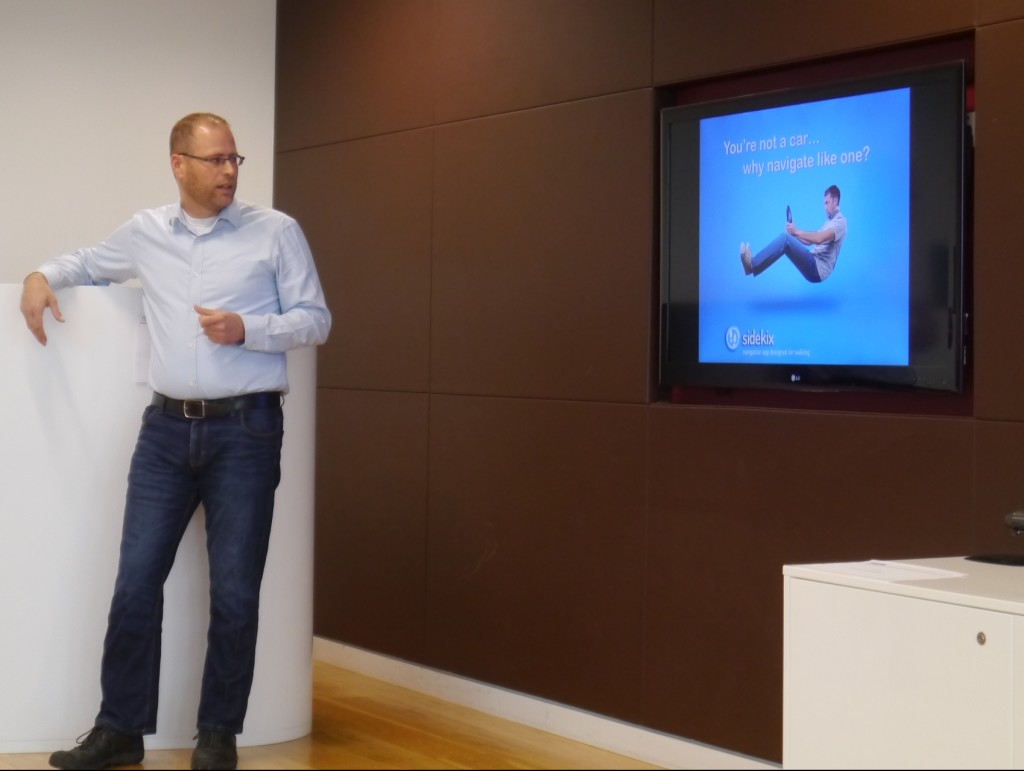 Sidekix 联合创始人 Miron Perel 站在演示屏幕旁边。上面是一个人漂浮在坐姿上的照片，好像在驾驶汽车，周围没有车辆。文字写道：“你不是汽车……为什么要像汽车一样导航？”